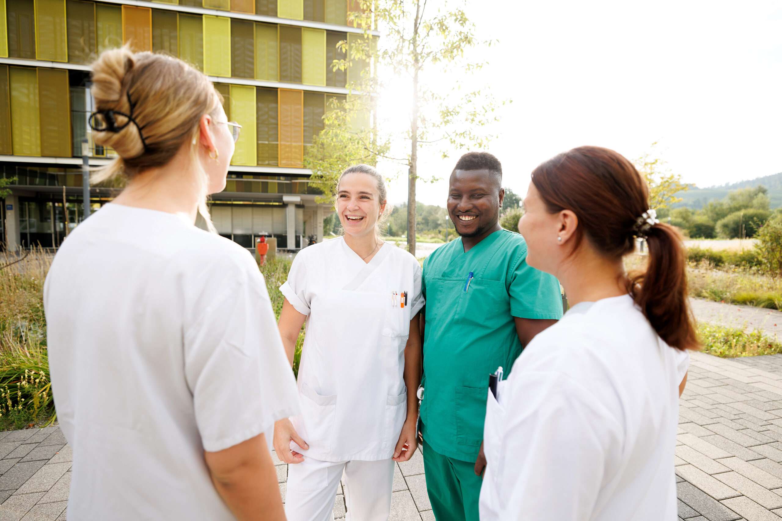 Drei Pflegefachfrauen und ein Pflegefachmann stehen vor dem Hauptgebäude Winnenden, sie sprechen miteinander und lachen